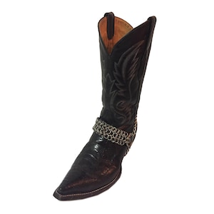 May include: Black cowboy boot with a pointed toe and decorative stitching. A silver chain boot strap wraps around the ankle. The boot has a textured leather appearance and a tall shaft.