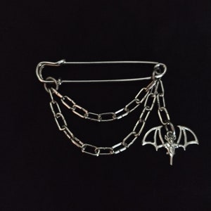 Puede incluir: Un broche de seguridad plateado con una cadena y un dije de murciélago. La cadena está hecha de tres hebras de eslabones plateados.