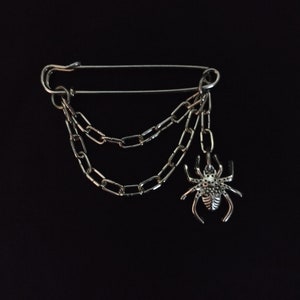 Peut inclure: Épingle de sûreté argentée avec trois chaînes pendantes et un charme araignée. L'araignée a des pattes et un corps détaillés. L'épingle est posée sur un fond noir.