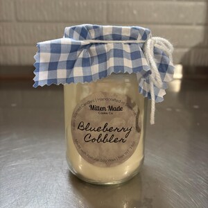 Pode incluir: Uma vela de vidro em um frasco com uma tampa de tecido xadrez azul e branco. O rótulo diz "Mitten Made Cookie Co. Blueberry Cobbler Made with Natural Soy Wax | Net WT 16oz."