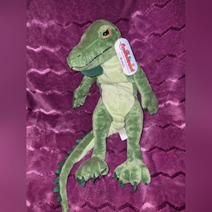 May include: A green plush alligator toy with a light green belly, dark green claws, and a long tail. The toy has a tag attached. The alligator is on a purple, textured surface.