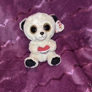 May include: A cream-coloured panda bear soft toy with black ears and feet, and large, glittery gold and black eyes. The panda holds a red heart with the word "Austria" printed on it. The background is a textured, deep purple fabric.