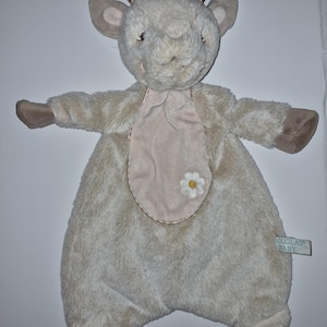 May include: A plush goat security blanket in light beige with brown striped horns and brown paws. The centre features a smooth, light pink oval patch with an embroidered white daisy flower. A small tag on the side reads Douglas Baby.