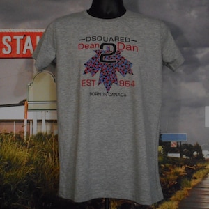 May include: A gray t-shirt with a crew neck featuring a colorful maple leaf design. The shirt has the text "DSQUARED Dean Dan" above the leaf and "EST 1964 Born in Canada" below it.