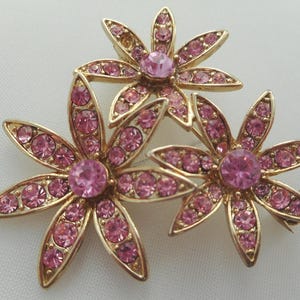 May include: A gold-toned metal brooch featuring three flower designs. Each flower has six petals, each petal is adorned with small pink crystals. The center of each flower has a larger pink crystal. The brooch is set against a white background.