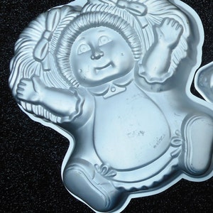 May include: White metal cake pan shaped like a doll with pigtails, wearing a dress and shoes.