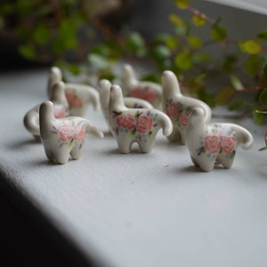 May include: A group of seven small white ceramic figurines of animals with pink floral designs. The figurines are arranged in a row on a white surface.