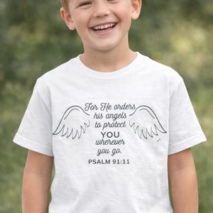 Puede incluir: Camiseta blanca con alas de ángel y el texto "For He orders his angels to protect YOU wherever you go. Psalm 91:11." Un niño sonriente lleva la camiseta.