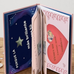 Könnte beinhalten: Offenes Buch mit rosa Einbänden und Seiten. Die linke Seite hat einen dunkelblauen Hintergrund mit einem Stern und spanischem Text. Die rechte Seite zeigt ein rotes Herz mit spanischem Text. Geld ist zwischen den Seiten sichtbar.