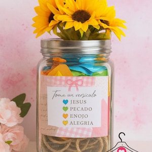 Puede incluir: Un frasco de vidrio lleno de papel de colores y cuerda, coronado con girasoles. Una etiqueta dice "Toma un versiculo" con palabras como "Jesús" y "Alegría". El frasco es un diseño de camiseta personalizada.