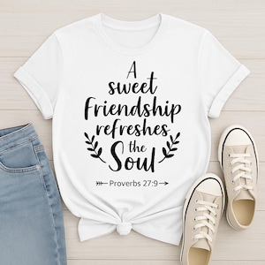 Puede incluir: Camiseta blanca con el texto "A sweet Friendship refreshes the Soul" en escritura negra. La camiseta tiene cuello redondo y mangas enrolladas. También se ven zapatillas de lona beige y vaqueros azules.