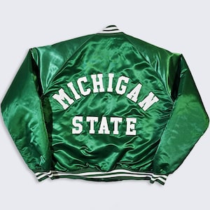 Puede incluir: Chaqueta de satén verde con las palabras "MICHIGAN STATE" en letras blancas en la espalda. La chaqueta tiene ribetes a rayas blancas y verdes en el cuello, los puños y la cintura. La chaqueta es de talla mediana.