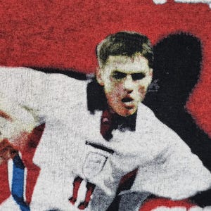 May include: A close-up of a man's face wearing a white jersey with a red and blue design. The man is looking directly at the camera with a serious expression. The background is red and black.
