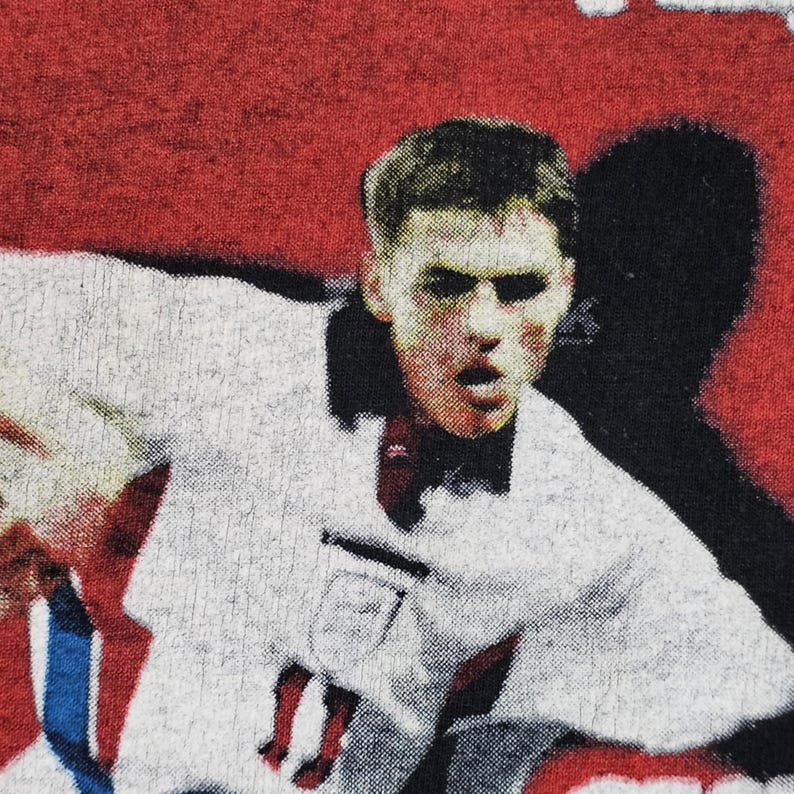 May include: A close-up of a man's face wearing a white jersey with a red and blue design. The man is looking directly at the camera with a serious expression. The background is red and black.