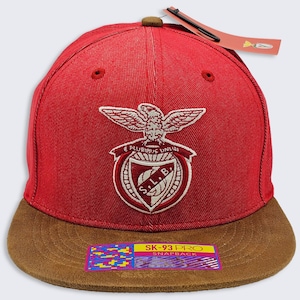 May include: A red baseball cap with a brown flat brim and a brown top button. The front features an embroidered crest with an eagle, the text E PLURIBUS UNUM, and S.L.B. A rectangular sticker on the brim reads SK-93 PRO SNAPBACK with a colourful pattern.