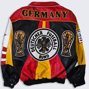 Könnte beinhalten: Eine schwarze, rote und gelbe Lederjacke mit dem Wort "GERMANY" auf dem Rücken. Die Jacke hat einen großen Patch mit den Worten "Deutscher Fussball Bund" und einem stilisierten Adler. Die Jacke hat auch zwei Patches einer Trophäe.