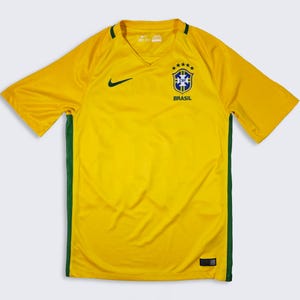 Puede incluir: Una camiseta de fútbol amarilla de Brasil con cuello en V y mangas cortas. Tiene una franja verde en los laterales y un logo de Nike. El escudo de la selección brasileña está en el pecho, con la palabra "BRASIL" debajo.
