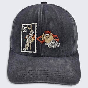 May include: A black baseball cap with an embroidered image of Bugs Bunny and Taz from Looney Tunes. The image of Bugs Bunny is on a postage stamp with the text "32 USA".