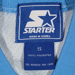 May include: A blue Starter brand tag with a white star and the text "Starter Made in Korea". Below the tag is a smaller blue tag with the text "S 100% Polyester See Reverse for Care".