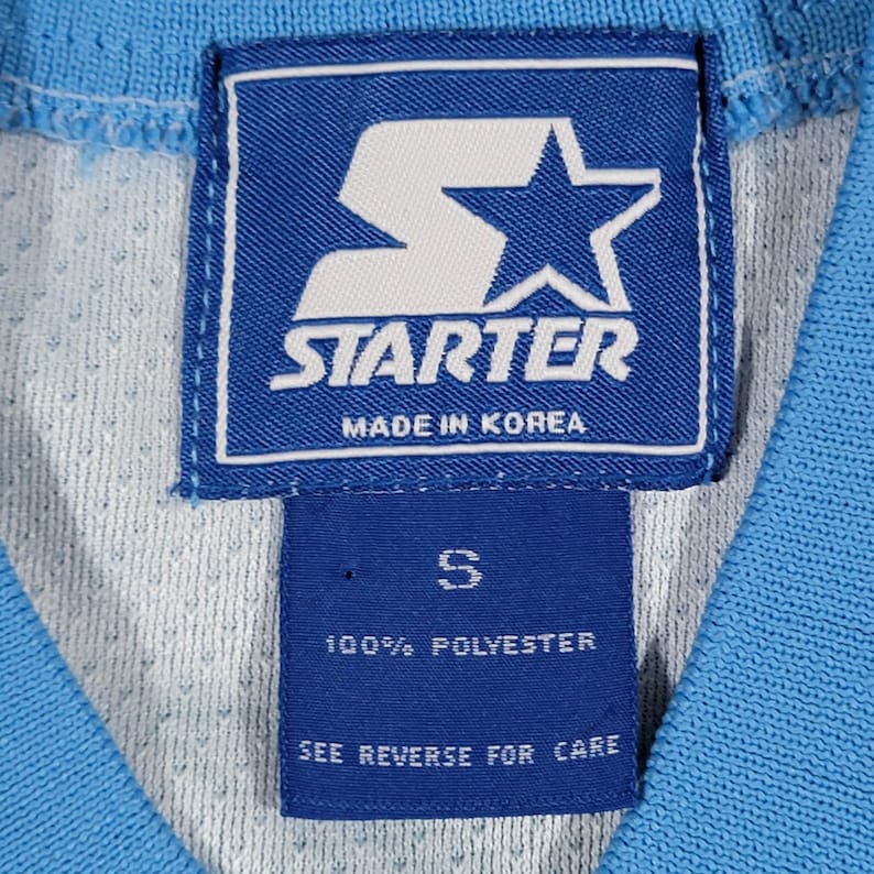 May include: A blue Starter brand tag with a white star and the text "Starter Made in Korea". Below the tag is a smaller blue tag with the text "S 100% Polyester See Reverse for Care".