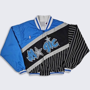 Op de afbeelding: Een blauwe, zwarte en witte satijnen baseballjas met het Orlando Magic-logo en "Magic" in grote witte letters. De jas heeft een gestreept patroon op de rechtermouw.