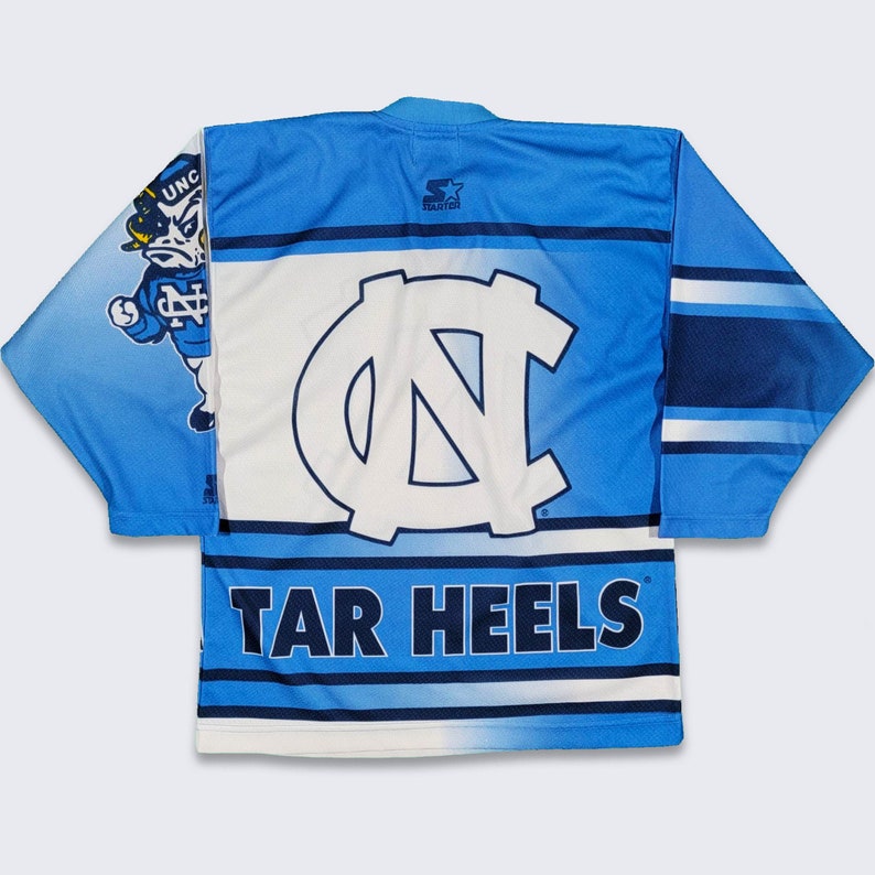 May include: A blue and white hockey jersey with the North Carolina Tar Heels logo and "TAR HEELS" text. The jersey has a blue and white striped pattern and features a cartoon mascot on the left sleeve.