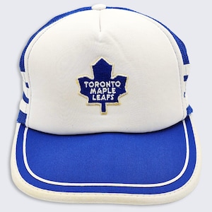 May include: A white and blue Toronto Maple Leafs baseball cap. The cap features a blue brim, white crown, and a blue and gold embroidered logo of a maple leaf with the text "TORONTO MAPLE LEAFS". The cap has a mesh back and a white button on top.