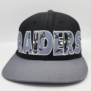 以下が含まれることがあります： 黒とグレーのベースボールキャップ。前面にグレーで「RAIDERS」と刺繍されています。レターにはレイダースのロゴが組み込まれています。