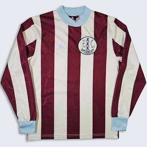 Puede incluir: Una camiseta de f&uacute;tbol de manga larga a rayas blancas y marrones con cuello azul. La camiseta tiene un logotipo en el pecho izquierdo y un parche en el pecho derecho que dice "F.C. Amsterdam".