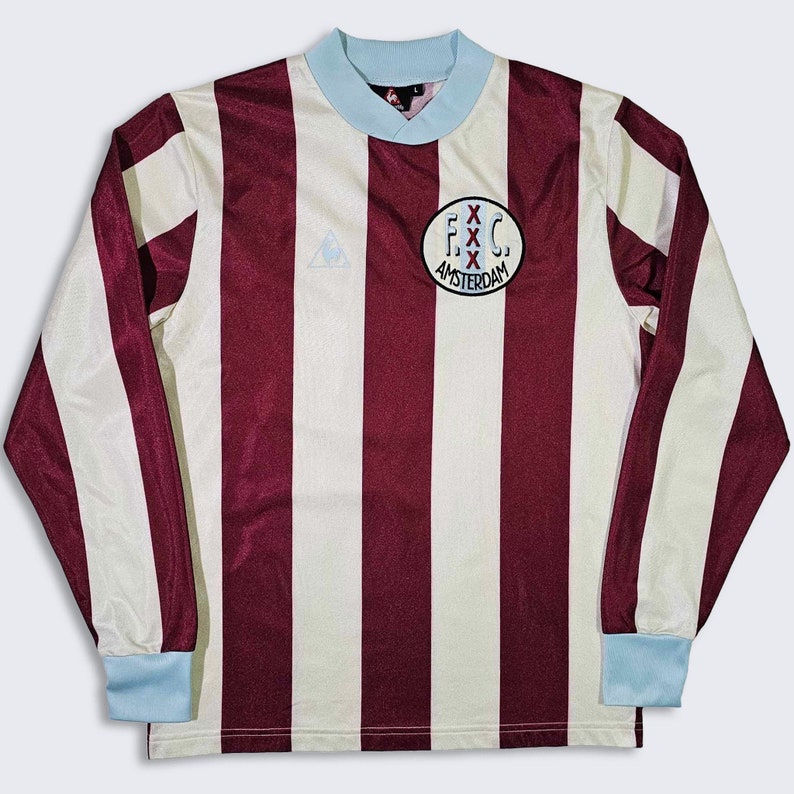 Puede incluir: Una camiseta de f&uacute;tbol de manga larga a rayas blancas y marrones con cuello azul. La camiseta tiene un logotipo en el pecho izquierdo y un parche en el pecho derecho que dice "F.C. Amsterdam".