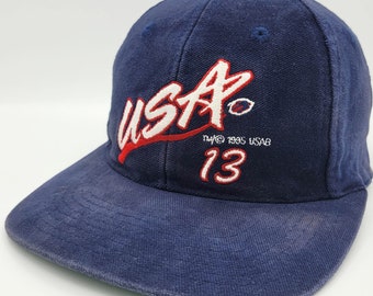 Op de afbeelding: Een marineblauwe baseballpet met een wit en rood "USA" logo en de tekst "TM 1995 USAG 13" geborduurd op de voorkant.