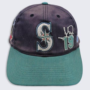 May include: A navy blue baseball cap with a teal brim. The front features an embroidered white and teal letter S with a compass star design, alongside an embroidered white signature and the number 19 in teal with a white outline.