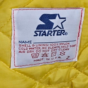 May include: A white tag with red and blue trim, featuring the Starter logo and care instructions. The tag reads "NAME SHELL & LINING: 100% NYLON COLD WATER, NO BLEACH, MILD SOAP AIR DRY. DO NOT DRY CLEAN MADE IN U.S.A. 2"