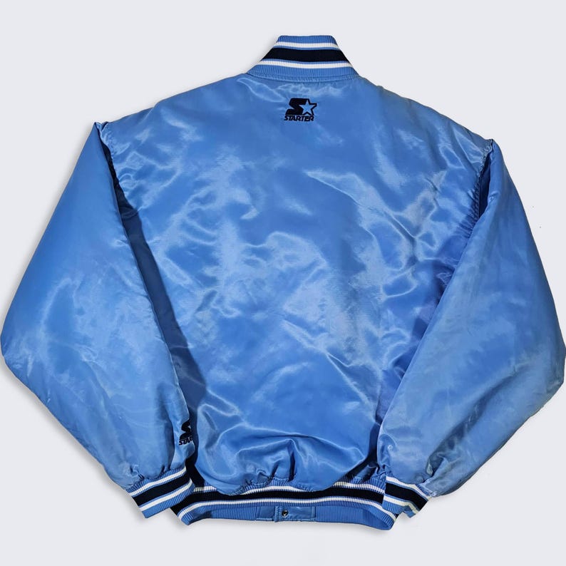 May include: Light blue satin jacket with black and white striped trim on the collar, cuffs, and hem. The Starter logo is visible on the upper back. The jacket has a snap closure.