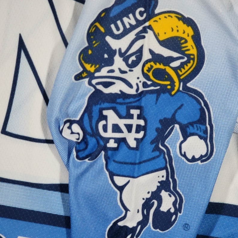 May include: A blue and white jersey featuring a cartoon ram mascot wearing a blue jersey with the letters "UNC" and "NC" on it.