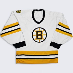 Puede incluir: Camiseta de hockey blanca con detalles en negro y dorado. La camiseta presenta un cuello negro, puños negros y rayas doradas y negras en las mangas. Un gran logotipo circular con una "B" negra dentro de un círculo dorado está en el pecho.