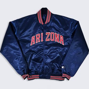 May include: A navy blue satin jacket with the word "ARI ZONA" in white and red lettering across the chest. The jacket has a red, white, and black striped collar, cuffs, and waistband. A small white star logo is visible on the sleeve.
