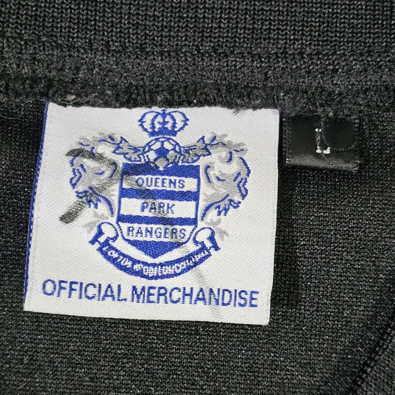 K&ouml;nnte beinhalten: Nahaufnahme eines schwarzen Kleidungsst&uuml;cks mit einem wei&szlig;en Etikett mit dem Logo der Queens Park Rangers und dem Text "OFFICIAL MERCHANDISE". Ein kleines schwarzes Etikett mit dem Buchstaben "L" ist ebenfalls sichtbar.