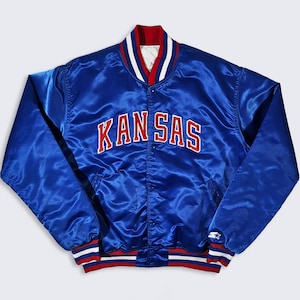 May include: A royal blue satin jacket with the word "KANSAS" in red block letters across the chest. The jacket has a red, white, and blue striped collar and cuffs. The Starter logo is visible on the lower right side.