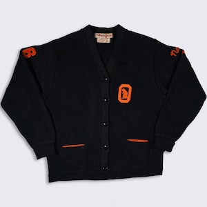 Puede incluir: Suéter de punto negro con ribetes y letras naranjas. El suéter tiene cuello en V, cierre de botones y dos bolsillos. Las letras en el suéter dicen "Newcomb" y "8".