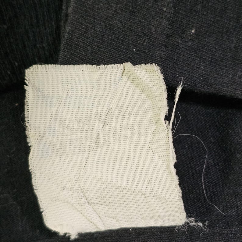 May include: A close-up of a white fabric label sewn onto a black fabric surface. The label is slightly wrinkled and has a rough texture.