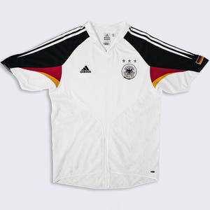 May include: White soccer jersey with black and red accents. Features the Adidas logo and the German national team emblem. The jersey has a V-neck and short sleeves.