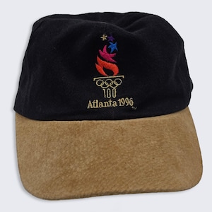 May include: A black baseball cap with a tan suede-like brim. The front features an embroidered design of a stylized torch with colorful flames above the Olympic rings, the number 100, and the text Atlanta 1996 in gold thread embroidery.