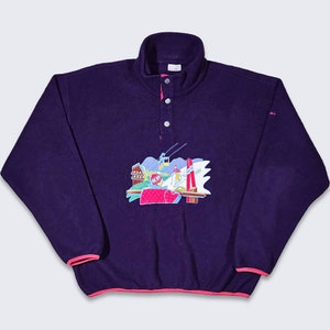 Puede incluir: Un jersey de forro polar morado con ribete rosa. El forro polar tiene un diseño bordado de colores de una estación de esquí con un telesilla, esquiadores y montañas.