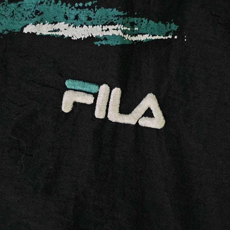 May include: Close-up of a black fabric with the white and teal FILA logo embroidered on it. The logo is in a bold, sans-serif font. Above the logo is a teal and white abstract design.