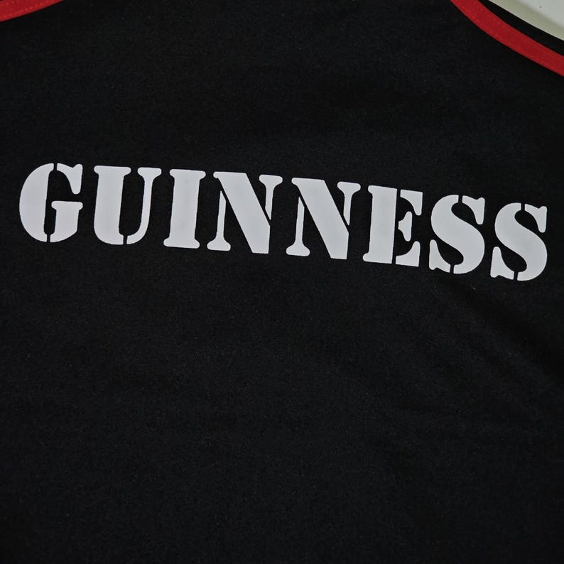 K&ouml;nnte beinhalten: Schwarzes Sport-T-Shirt mit dem Wort "GUINNESS" in gro&szlig;en wei&szlig;en Schablonenbuchstaben auf der Vorderseite. Das Shirt hat einen rot-wei&szlig;en Besatz am Halsausschnitt.