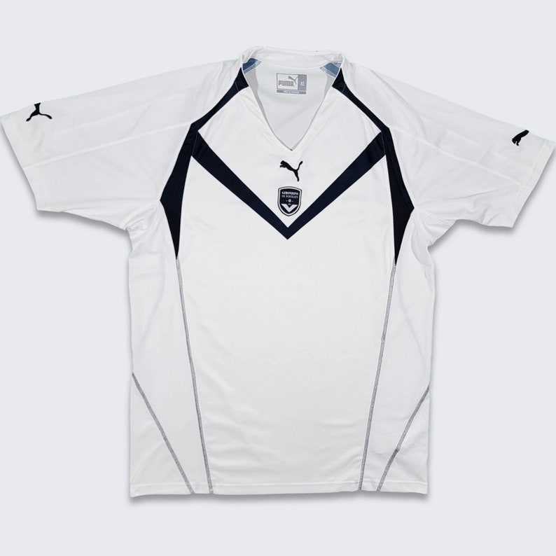 K&ouml;nnte beinhalten: Wei&szlig;es Fu&szlig;balltrikot mit schwarzen und blauen Akzenten. Das Trikot hat einen V-Ausschnitt und das Puma-Logo auf der Brust. Das Arsenal-Wappen befindet sich ebenfalls auf der Brust.