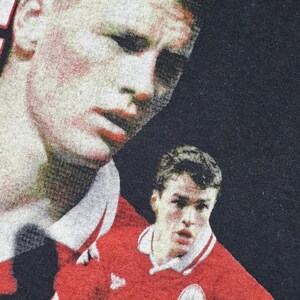 May include: A black t-shirt with a printed image of two soccer players in red jerseys. The players are looking at the camera. The shirt has a vintage look.