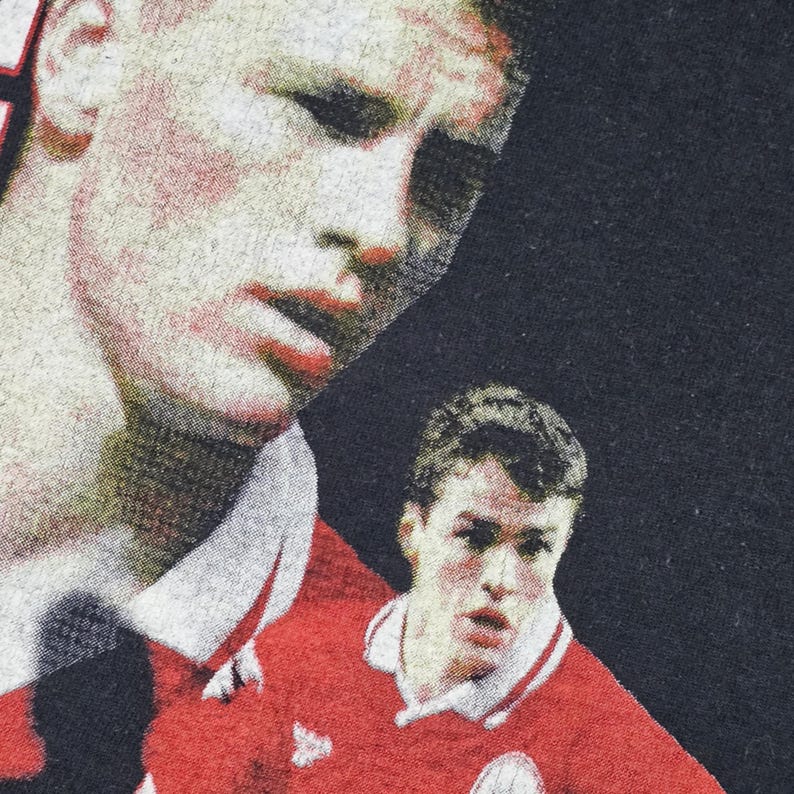 May include: A black t-shirt with a printed image of two soccer players in red jerseys. The players are looking at the camera. The shirt has a vintage look.
