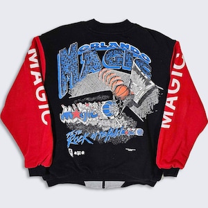 Puede incluir: Sudadera negra con mangas rojas y un estampado gráfico de un jugador de baloncesto haciendo un mate. El gráfico incluye el texto "Orlando Magic" y "Put the Rock in the Hole".